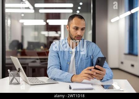 Latin American young man businessman, freelancer working in the office, sitting at the table, seriously and concentratedly using the mobile phone. Stock Photo