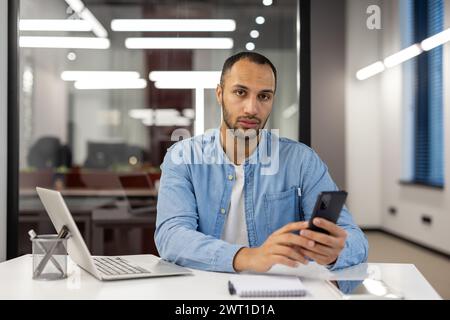 Portrait of a serious Latin American man sitting in the office at the desk, holding a telephone in his hands and looking at the camera. Stock Photo