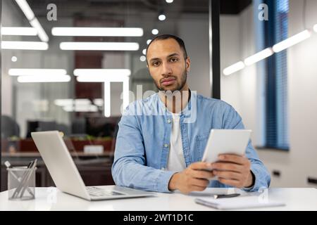 Portrait of a young serious Muslim man sitting in the office at a desk with a notebook and documents, holding a tablet in his hands and looking seriously into the camera. Stock Photo