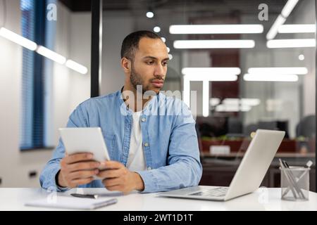 A serious young man, a dancer, a designer works in the office on a laptop, sits at a table and uses a tablet in his hands. Stock Photo