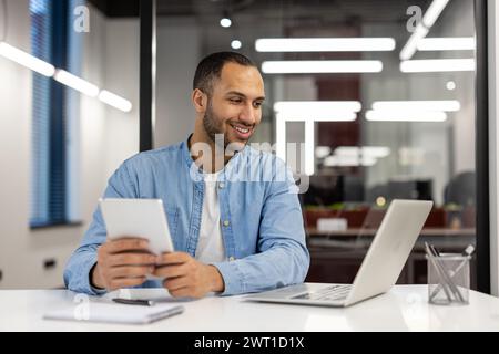 A young Hispanic man is talking on a video call on a laptop, sitting at a desk in the office, looking at the screen with a smile and holding a tablet in his hands. Stock Photo
