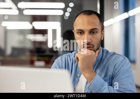 A close-up portrait of a young African American man sitting in the office in front of a laptop screen, intently and focused on work, a project, thinking about a solution to a problem. Stock Photo