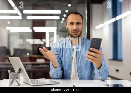 Serious and worried young man sitting in the office at the desk, holding a mobile phone in his hand and looking disappointedly at the camera while spreading his hands. Stock Photo