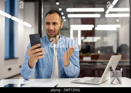 A young African American man works in the office sitting worried at the desk and looking at the phone screen in frustration and shock. Stock Photo