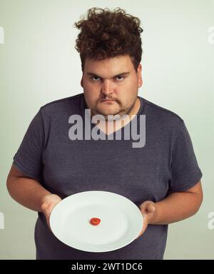 Portrait, diet or plate and plus size man in studio on gray background, unhappy with size of meal. Food, health or nutrition and disappointed young Stock Photo