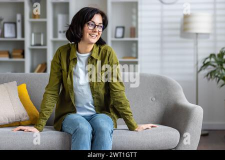 Smiling young woman wearing casual clothes and glasses sitting relaxed on sofa at home, looking away. Stock Photo