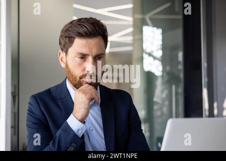 Close-up photo of a young serious male businessman sitting focused in the office at a laptop and thinking about information, leaning his head on his hand. Stock Photo