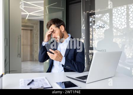 Shocked young man businessman sitting in the office on a chair and holding his head with his hand, holding the phone, upset by the news he received. Stock Photo