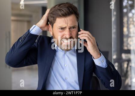 Close-up photo of a young businessman man in a suit standing in an office center, worriedly talking on the phone and holding his head with his hand in frustration, solving financial problems. Stock Photo