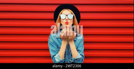 Portrait of stylish young woman model blowing her lips sending sweet air kiss wearing jean jacket, black round hat on red background Stock Photo