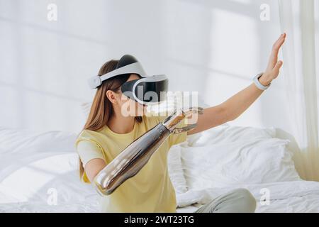 A woman explores virtual reality, wearing a VR headset and interacting with a futuristic robotic arm in a bright bedroom, symbolizing the blend of virtual experiences with advanced technology. Woman Experiencing VR with Robotic Arm Stock Photo