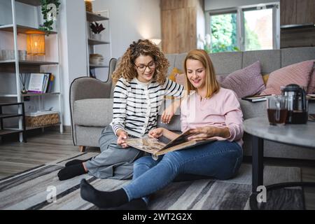 Two female friends are sitting on the floor in the living room, smiling and looking at and flipping through an old photo album. Stock Photo