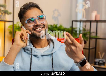 Phone call, good news, gossip. Portrait of happy Indian man having pleasant conversation on smartphone, enjoying talking with friend at modern home room apartment. Guy enjoying mobile phone speaking Stock Photo