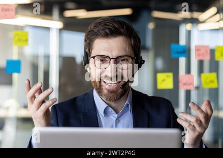 A happy, bearded businessman in a blue suit using a laptop. He wears a headset, gesturing during a video call in a modern office with sticky notes on the glass. Stock Photo