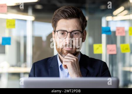 Focused male professional using laptop at a workspace with sticky notes in the background for brainstorming. Stock Photo