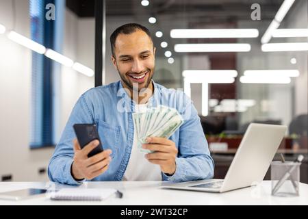 Happy businessman in a casual blue shirt holding cash and a mobile phone, feeling successful at work in a modern office. Stock Photo