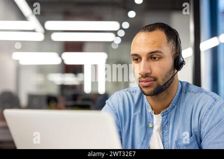 A concentrated male worker wearing a headset while being engaged with tasks on his laptop in a modern office setting. Stock Photo
