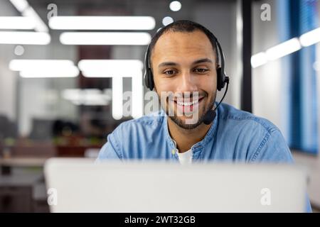 A cheerful male customer service representative wearing a headset with a microphone, working on a computer in a contemporary office setup. Stock Photo