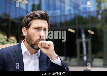 Close-up photo of a young sick man in a business suit who is standing outside an office building and coughing, covering his mouth with his hand. Stock Photo