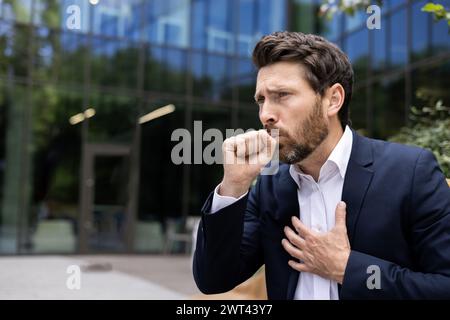 A young male businessman suffers from asthma and allergies. He sits outside an office building on a bench, coughs covering his mouth with his hand and holds his chest in pain. Stock Photo