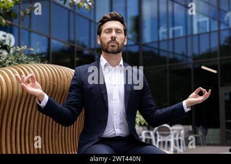 Relaxed young male businessman sitting on bench near office center wearing suit and lotus yoga pose, eyes closed, meditating and resting on break. Stock Photo