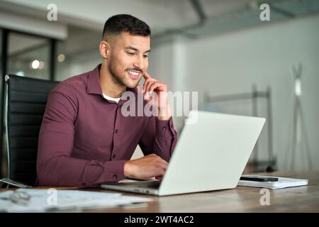 Happy busy Latin business man working on laptop sitting at desk in office. Stock Photo