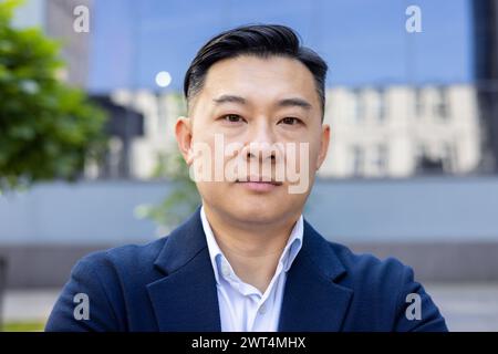 Confident Asian male executive in a stylish blue suit and white shirt, looking seriously at the camera outdoors. Stock Photo