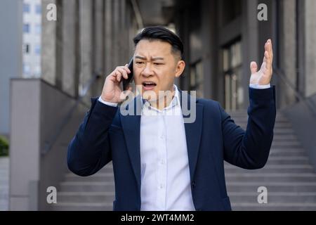 An upset executive on a stressful phone call, expressing frustration while standing outside office buildings in an urban setting. Stock Photo
