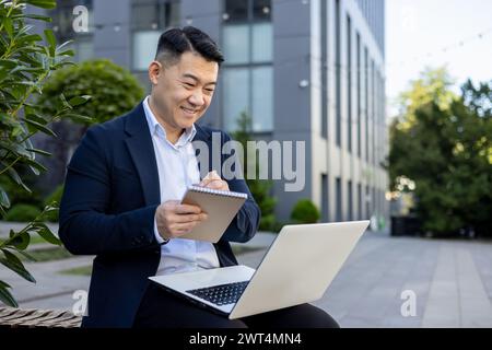 Cheerful Asian male professional taking notes and using laptop while working outside office premises. Stock Photo
