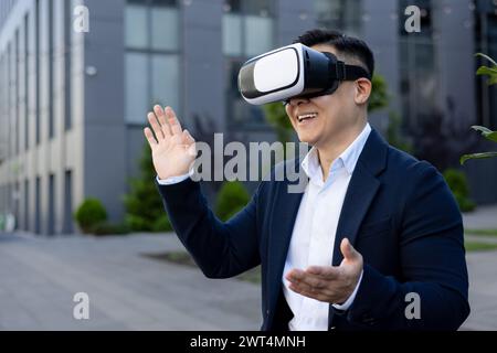 Enthusiastic businessman in a suit interacts with virtual reality, smiling and waving against an urban backdrop. Stock Photo