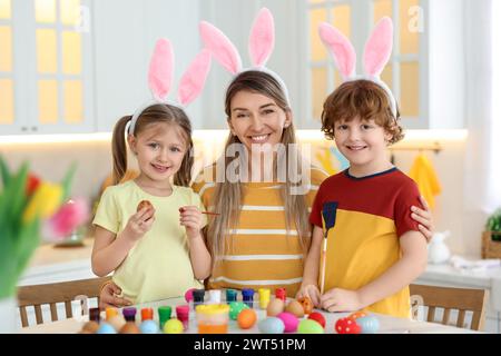 Painting Easter eggs. Happy mother and her cute children with bunny ears at table in kitchen Stock Photo