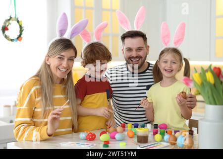 Painting Easter eggs. Happy family with bunny ears at white marble table in kitchen Stock Photo