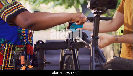 Healthy african american girlfriend assisting her caucasian boyfriend fix his bicycle outside. Active young woman fastening bike to repair-stand for athletic man to repair in home yard, closeup. Stock Photo