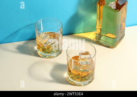 Whiskey with ice cubes in glasses and bottle on white table against light blue background Stock Photo