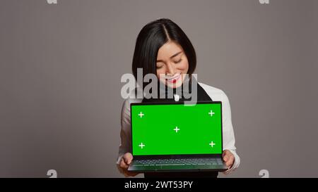 Asian server presenting greenscreen display on laptop in studio, wearing uniform with apron and bow. Restaurant hostess holding pc with blank mockup template, copyspace layout. Camera B. Stock Photo