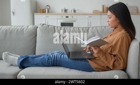 Side view Asian woman girl on sofa with laptop student writing notes in copybook preparing for exam doing homework working from home watching Stock Photo