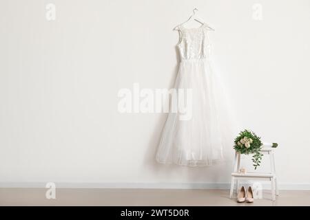 Beautiful wedding dress hanging on white wall, wedding shoes and bridal bouquet in room Stock Photo