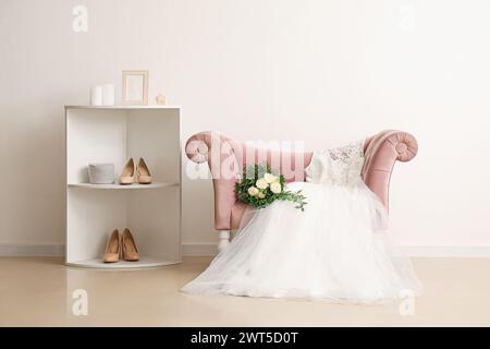 Wedding dress, bridal bouquet on pink armchair and wedding shoes on shelf near white wall in room Stock Photo