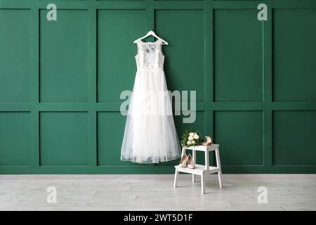 Beautiful wedding dress on green wall with ring box, wedding shoes and bridal bouquet on white wooden stepladder Stock Photo