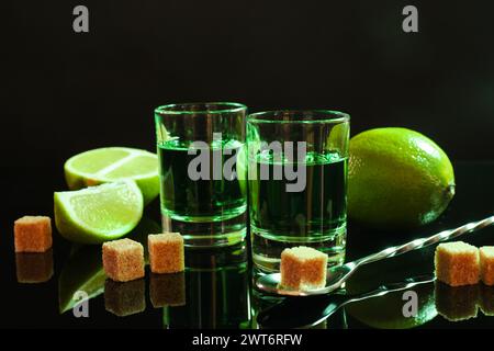 Absinthe in shot glasses, brown sugar, lime and spoon on mirror table. Alcoholic drink Stock Photo