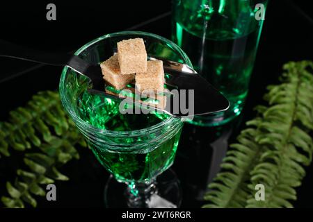 Absinthe in glass, brown sugar, spoon and green leaves on mirror table, closeup. Alcoholic drink Stock Photo