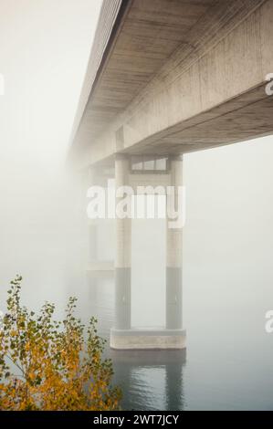 Concrete bridge disappearing in a dense fog. Bridge over a river - low visibility on November morning. Perspective view of the bridge from river bank. Stock Photo
