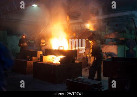 Workers in protective gear operate in a foundry, handling molten metal with machinery. Industrial manufacturing process, metallurgy tools in use Stock Photo