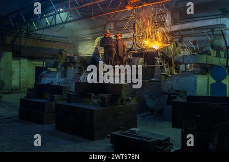 Industrial workers in protective gear casting metal in foundry. Team supervises molten steel pouring, safety helmets, heavy machinery. Manufacture Stock Photo
