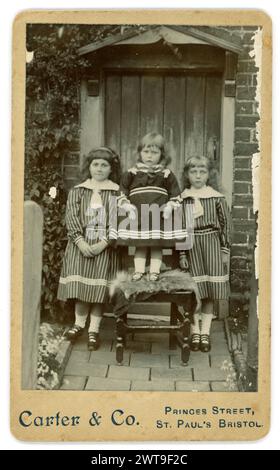 Original Victorian carte de visite (visiting card or CDV) 3 Victorian children outside the front door in the garden, in summer, wearing striped dresses with sailor suit features, popular at the time, white socks and strappy sandals.  From the studio of Carter & Co. Princes Street, St. Paul's, Bristol. Circa 1885. Stock Photo