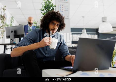 Young arab comapny employee drinking coffee and using corporate laptop in business office. Executive manager holding tea mug and solving work tasks on computer in coworking space Stock Photo