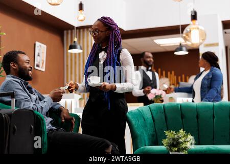 Hotel waitress holding cup of freshly brewed coffee taking cash payment from guest while serving hot drink. Happy African American man traveler ordering espresso while waiting in lobby for check-in Stock Photo