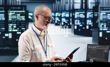 Meticulous computer scientist in data center, solving tasks on tablet while running scripts on laptop terminal. Precise admin amidst servers, accessing databases and manipulating lines of code Stock Photo