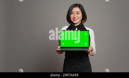 Asian server presenting greenscreen display on laptop in studio, wearing uniform with apron and bow. Restaurant hostess holding pc with blank mockup template, copyspace layout. Camera A. Stock Photo