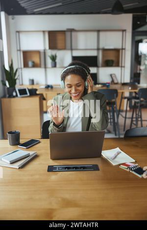 A woman wearing headphones sits at a desk with a laptop computer in nice modern coworking office Stock Photo